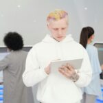 A young man with colorful hair and tattoos using a tablet in a modern tech environment while others work in the background.