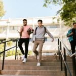 Three college students walk down stairs on campus, carrying books and backpacks, on a sunny day.
