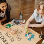 Two teenage girls painting a poster for an art project, promoting recycling and creativity. Indoors setting.