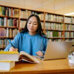 Young woman studying in a library with books and laptop, focusing on education.